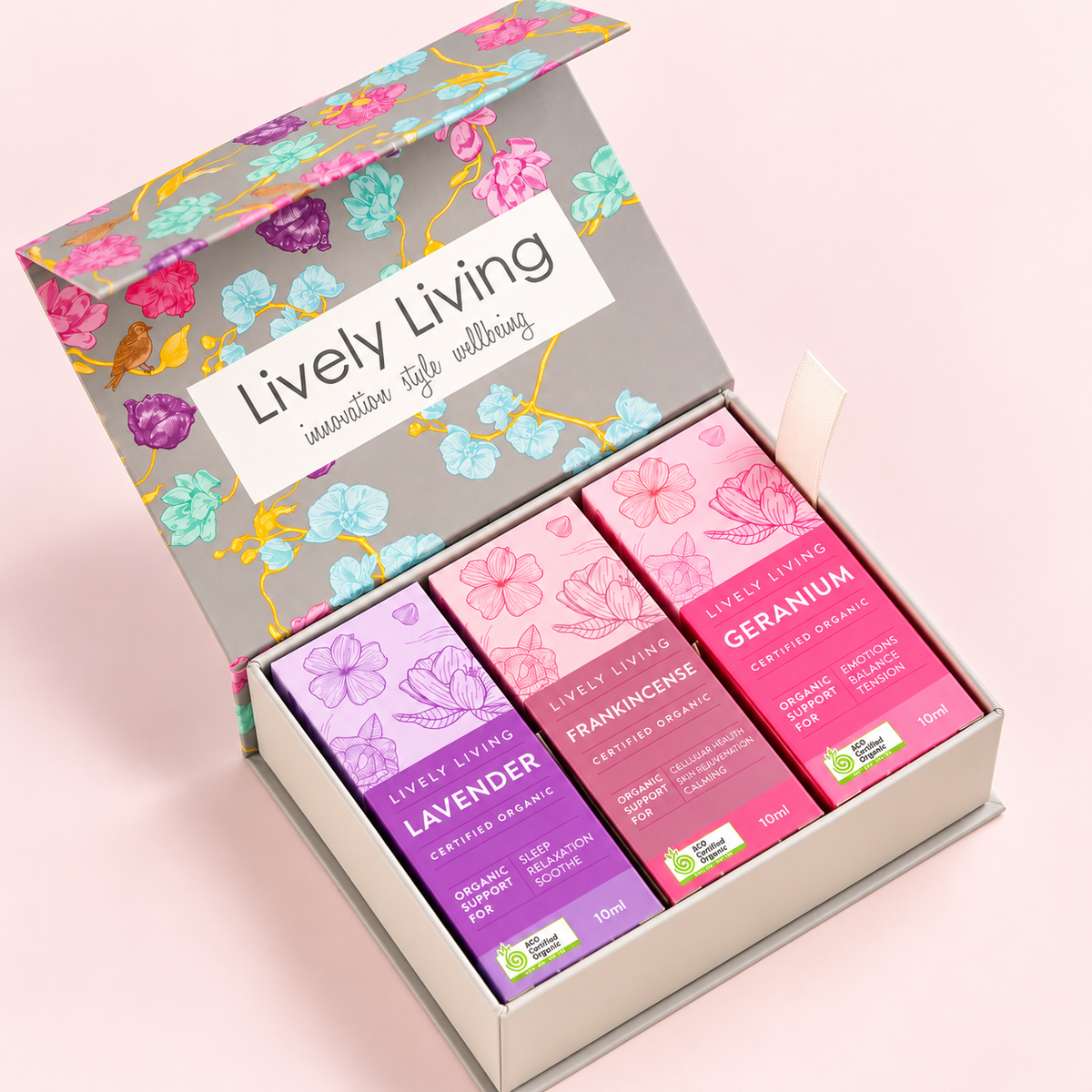 An open gift box containing four small herbal cream bars in different colors: lavender purple, frangincense pink, and geranium magenta. The box lid features a floral pattern with the brand name 'Lively Living' and the tagline 'innovative self wellness'.