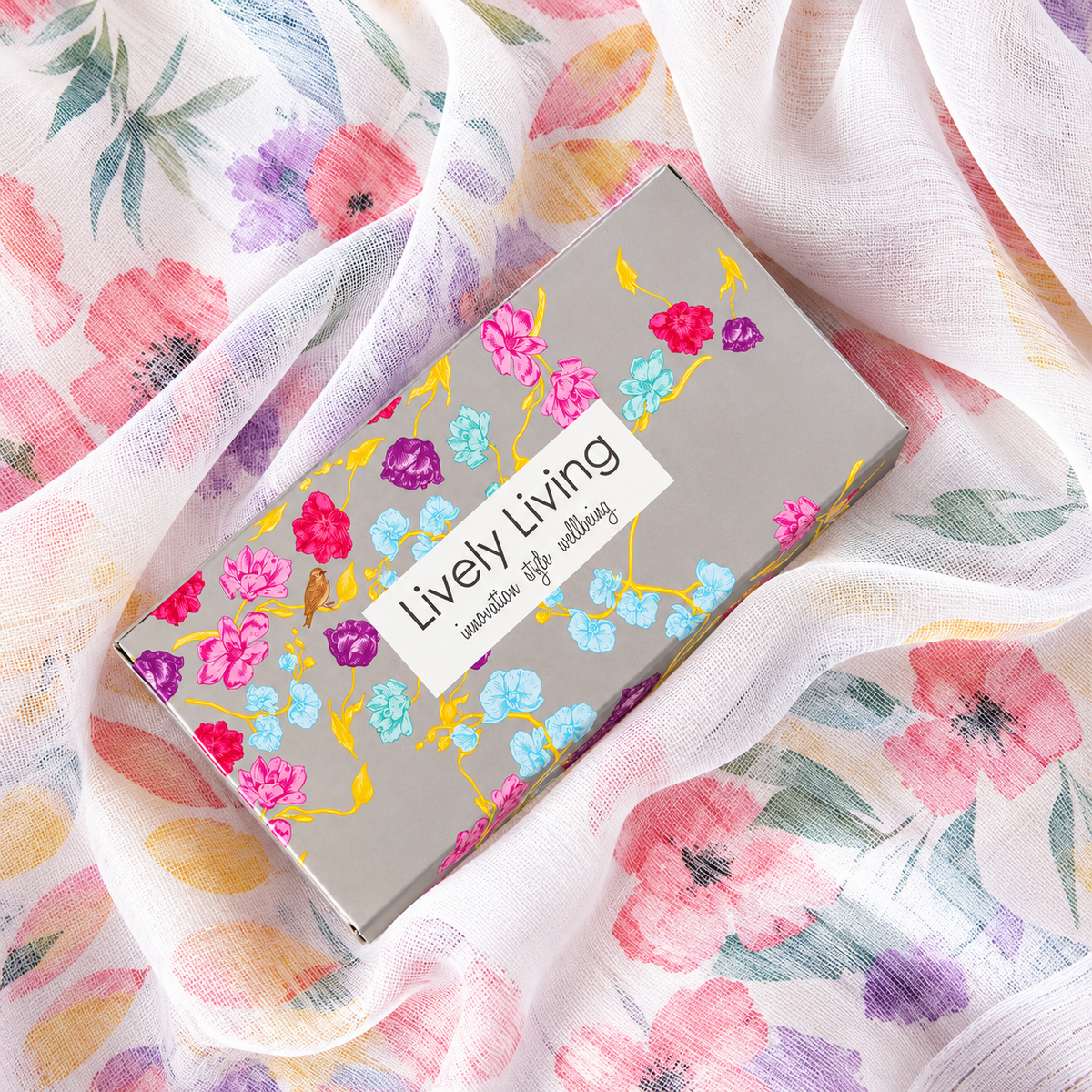 Closed grey Lively Living gift box printed with pink, purple and blue flowers, resting on sheer white floral fabric