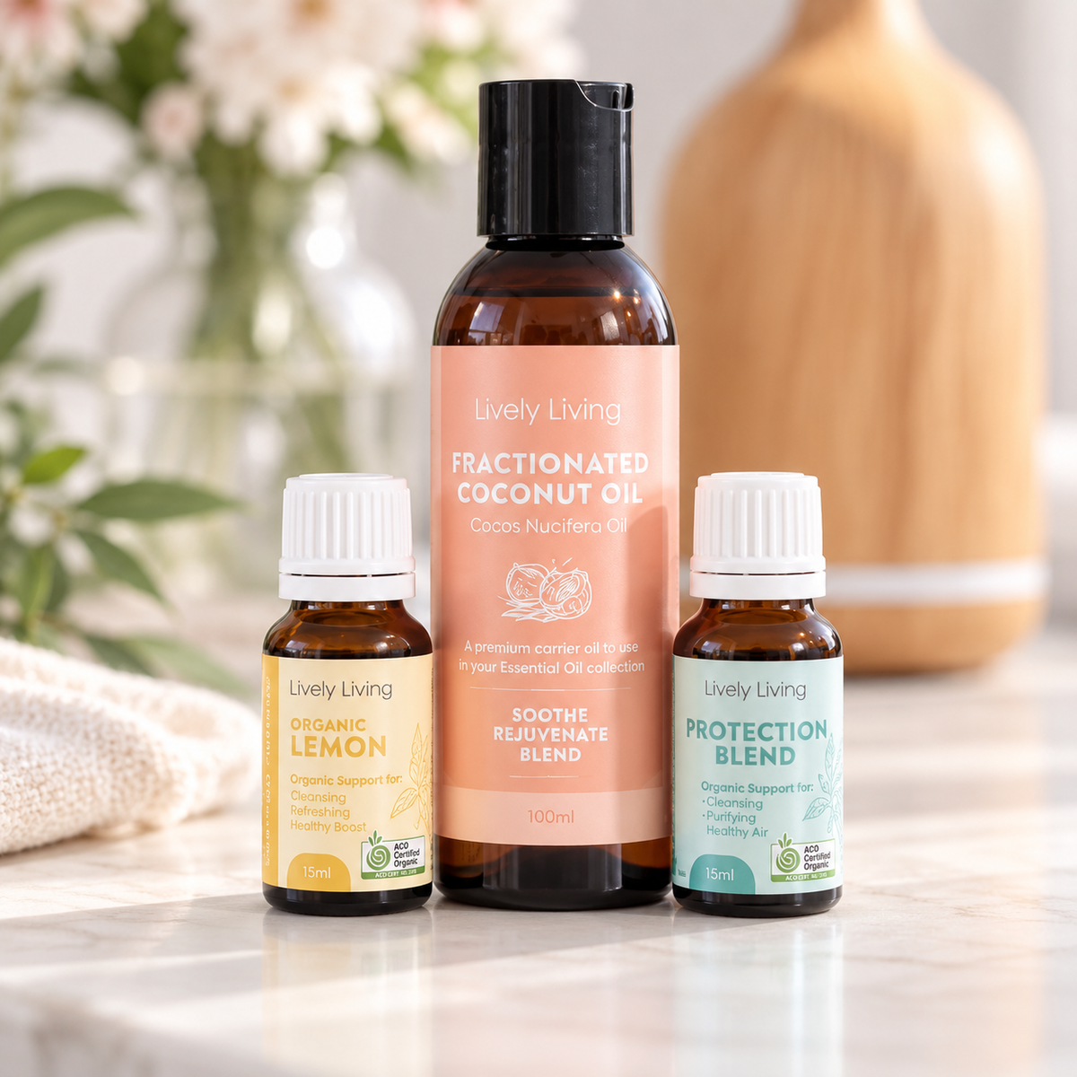 Lively Living Fractionated Coconut Oil bottle between Organic Lemon and Protection Blend essential oils on a marble bench, with a wood-look diffuser and white flowers behind