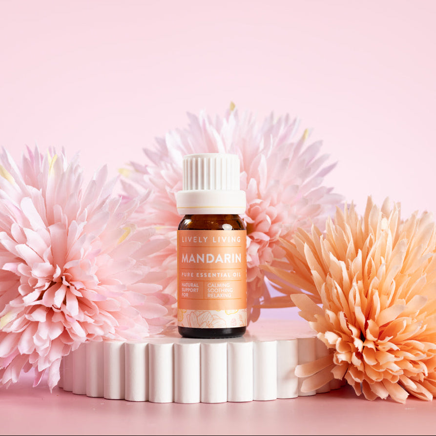 Lively Living Pure essential Mandarin Essential Oil, sitting on a white rippled plinth surrounded by pink and orange flowers. 