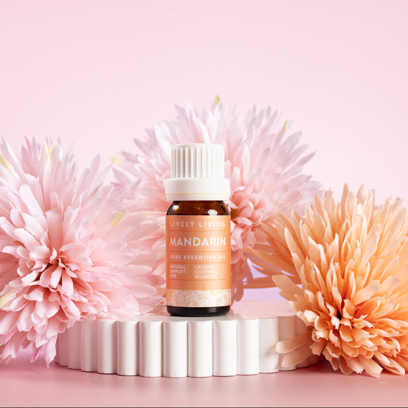 Lively Living Pure essential Mandarin Essential Oil, sitting on a white rippled plinth surrounded by pink and orange flowers. 