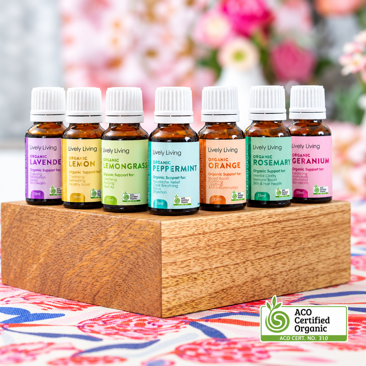 Six small amber glass essential oil bottles with white caps are arranged on a wooden block. The bottles have colorful labels in purple, yellow, light green, blue, orange, and pink, with the brand name 'Lively Living' visible on each.