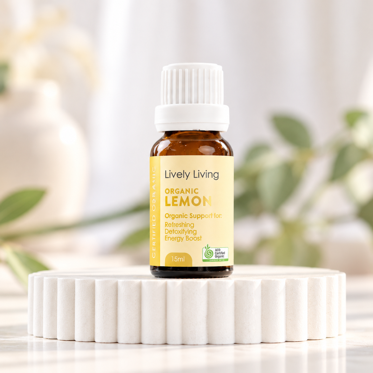 Lively Living Organic Lemon 15ml essential oil with a yellow label on a white fluted pedestal, with a white vase and green leaves behind