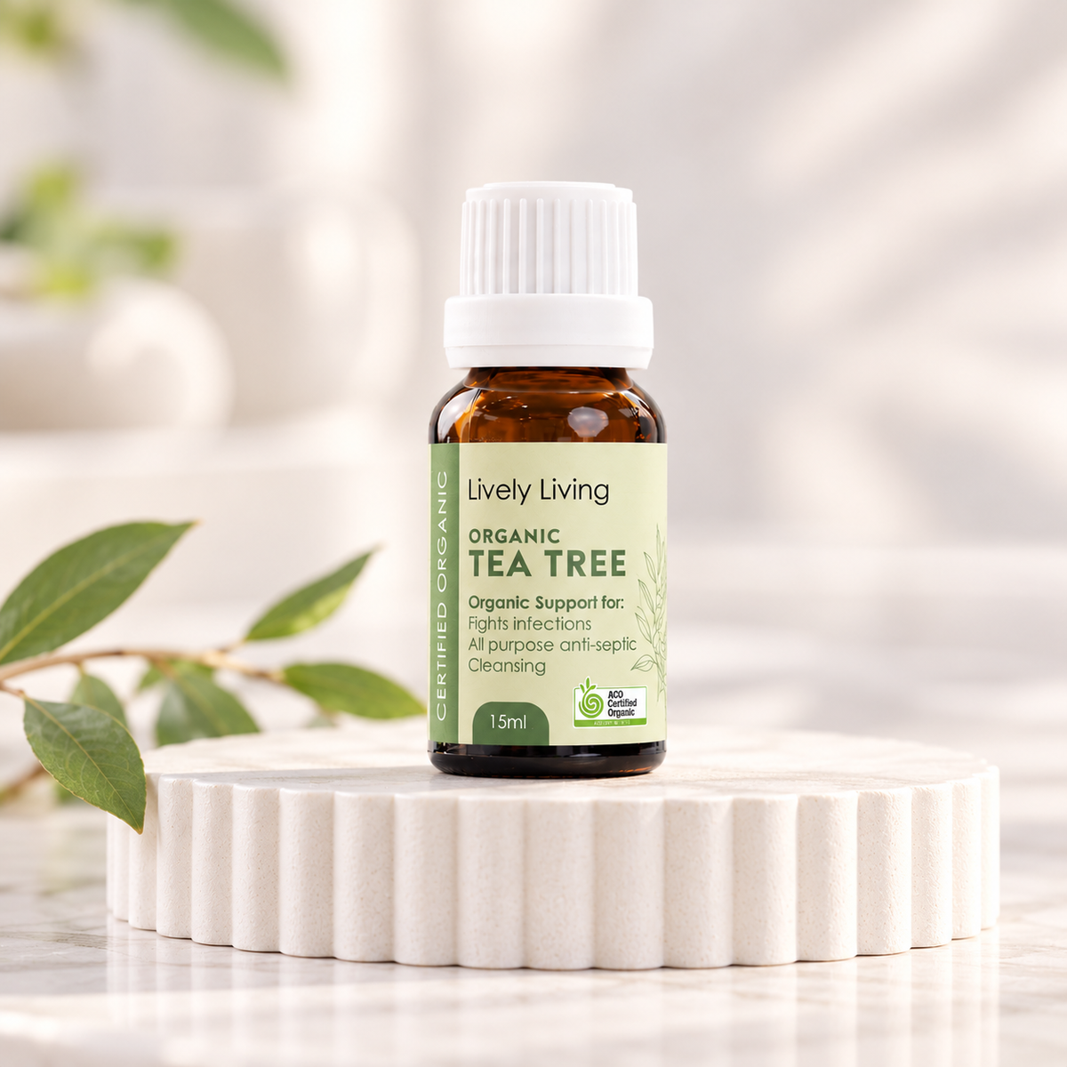 Lively Living Organic Tea Tree 15ml essential oil with a green label on a white fluted pedestal, with a sprig of green leaves beside it