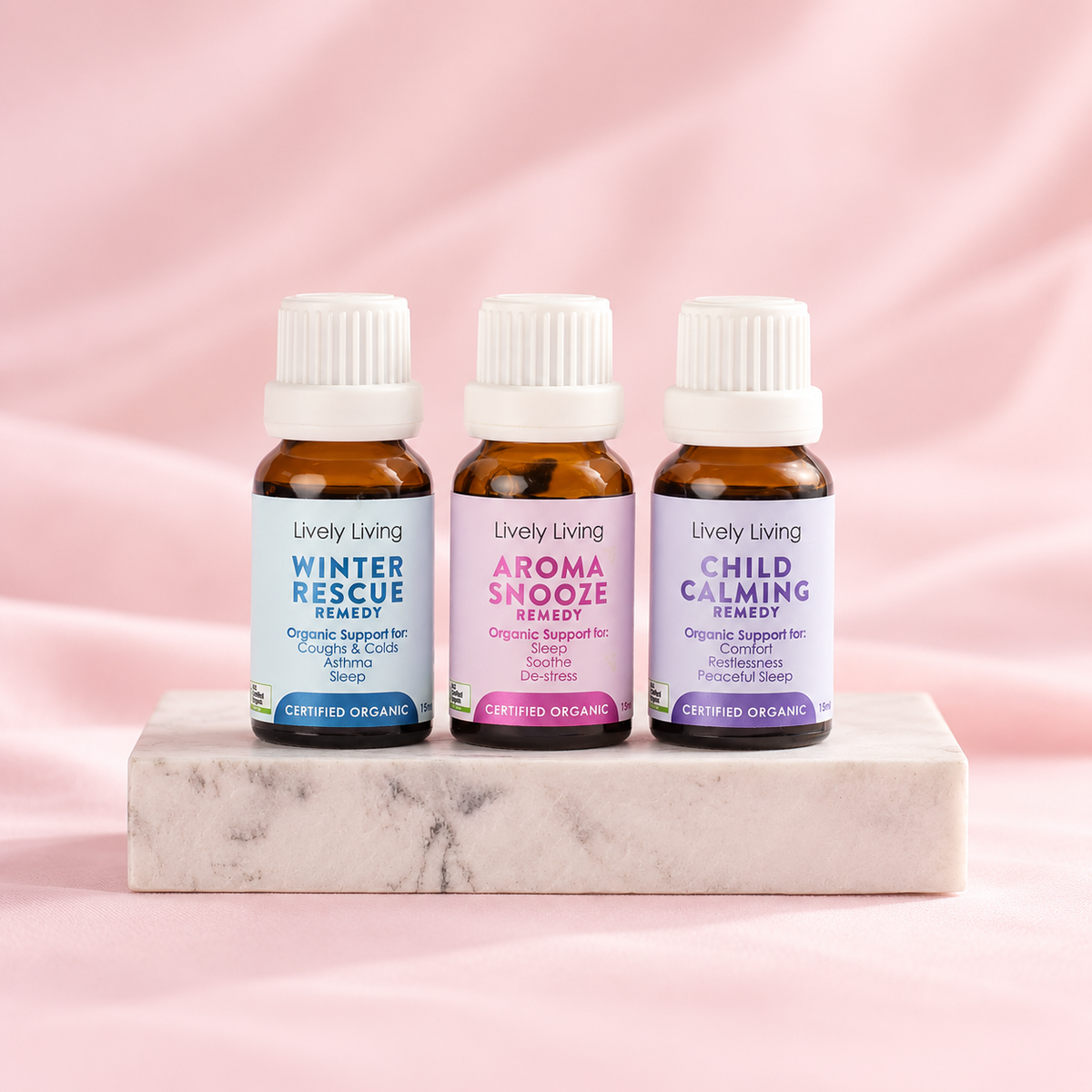Three Lively Living certified organic remedy oils, Winter Rescue, Aroma Snooze and Child Calming, in 15ml amber bottles on a marble block against pink fabric