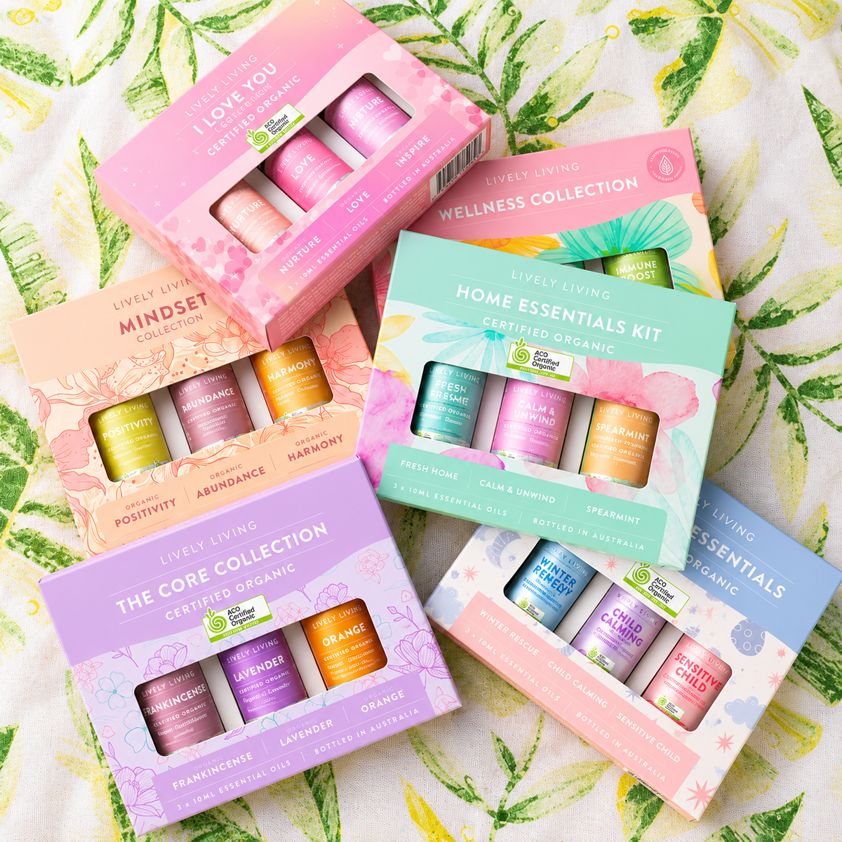 Flat lay of six Lively Living essential oil gift sets, including Mindset, The Core Collection and Home Essentials Kit, on palm leaf fabric