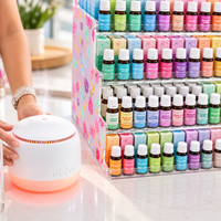 Hand resting on a white diffuser with a soft orange glow beside a tiered acrylic display stand filled with Lively Living essential oil bottles and boxes in pastel colours