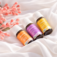 Three small essential oil bottles labeled Orange, Lavender, and Lemon, arranged on white draped fabric with pink decorative stems. The bottles have white caps and amber glass, with colorful labels in orange, purple, and yellow.