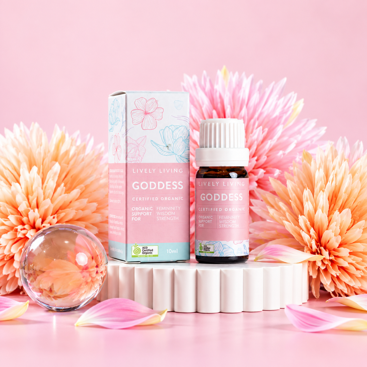 Goddess certified organic essential oil bottle and box on a white pedestal with pink and peach blooms and a glass sphere