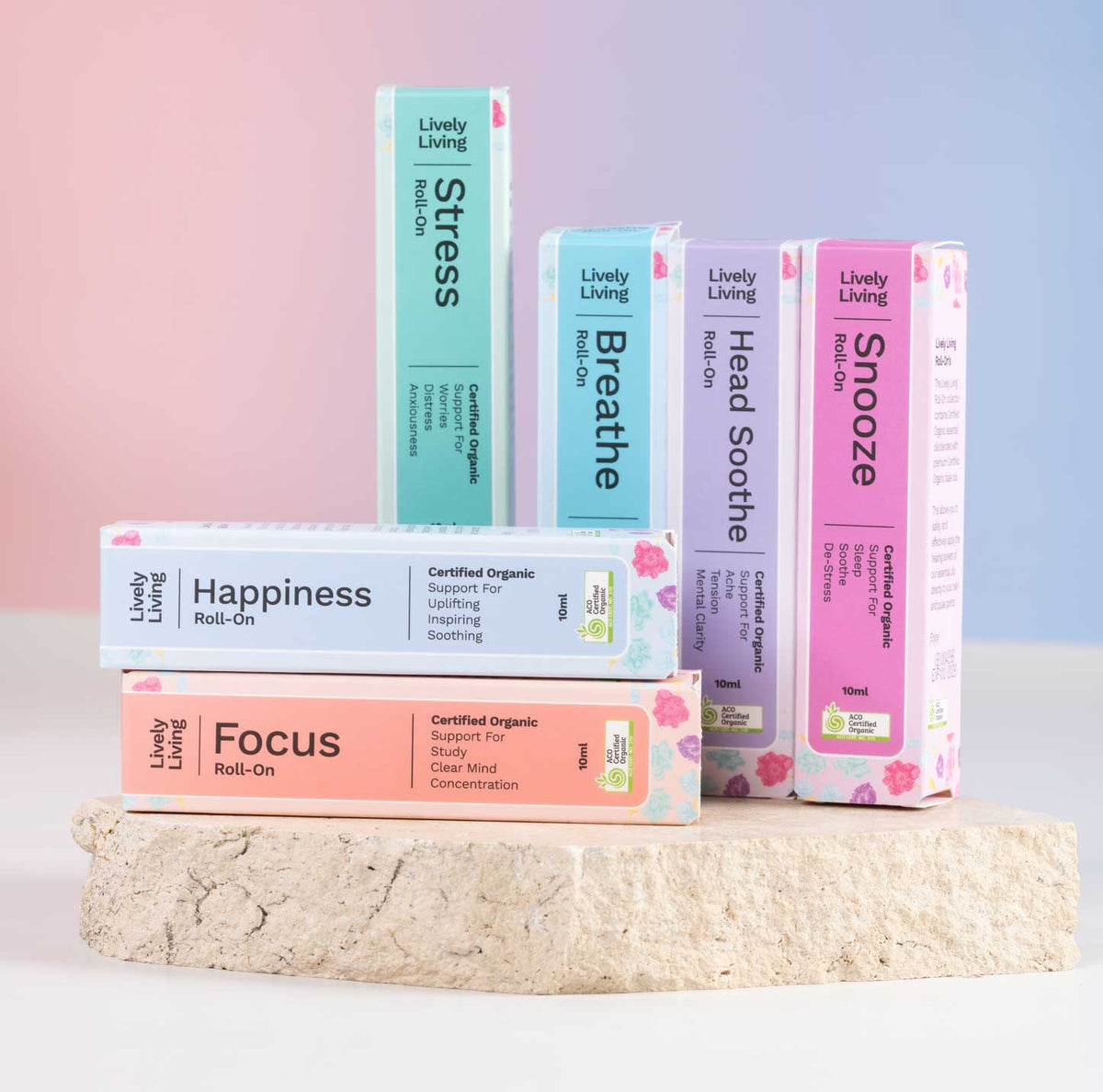 Six boxed wellness products from Lively Living are arranged on a light stone pedestal against a soft pink-to-blue gradient background. The boxes are labeled Stress, Happiness, Breath, Head Soothe, Focus, and Snoone, with pastel colors and small pink floral accents.