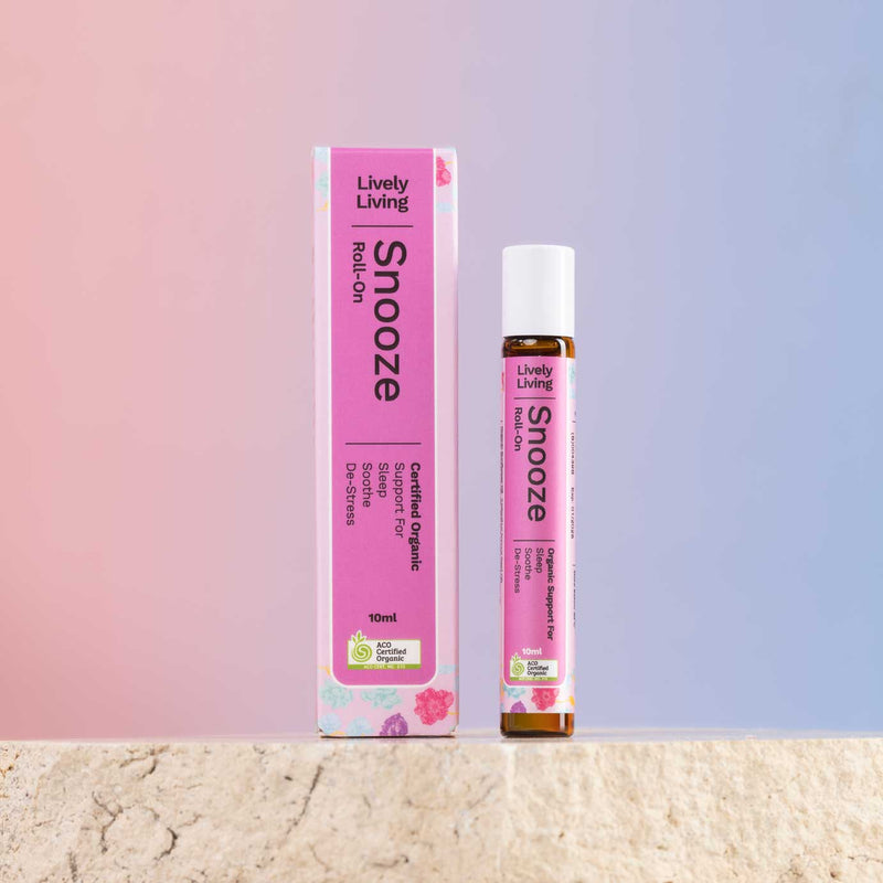 A pink roll-on deodorant bottle and its matching pink box from Lively Living, labeled 'Snooze', standing on a light stone surface against a soft pink-to-blue gradient background.