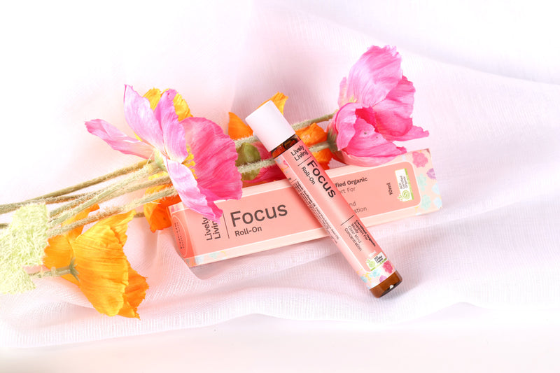 Living Living "Focus" roll on laying diaognal to the branded box next to faux flowers that are pink, on a sheet of sheer material