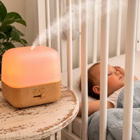 A glowing essential oil diffuser with a wood-grain base sits on a distressed wooden table beside a crib, emitting a soft mist. A sleeping baby is visible inside the crib, creating a cozy, serene nursery scene.