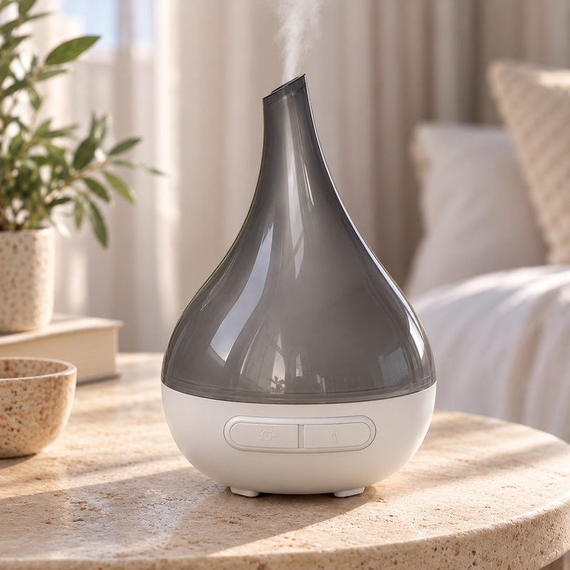 A teardrop-shaped essential oil diffuser with a glossy dark gray upper section and a white base, shown emitting a fine mist in a softly lit bedroom setting.