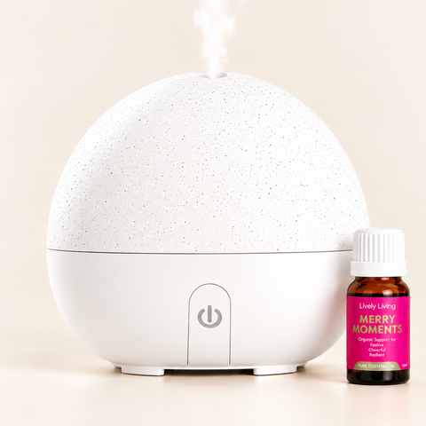 A white speckled spherical diffuser with mist coming from the top, shown next to a small amber bottle labeled 'Lively Living Merry Moment' on a warm beige background.
