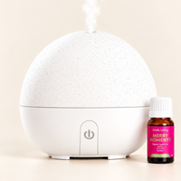 A white speckled spherical diffuser with mist coming from the top, shown next to a small amber bottle labeled 'Lively Living Merry Moment' on a warm beige background.