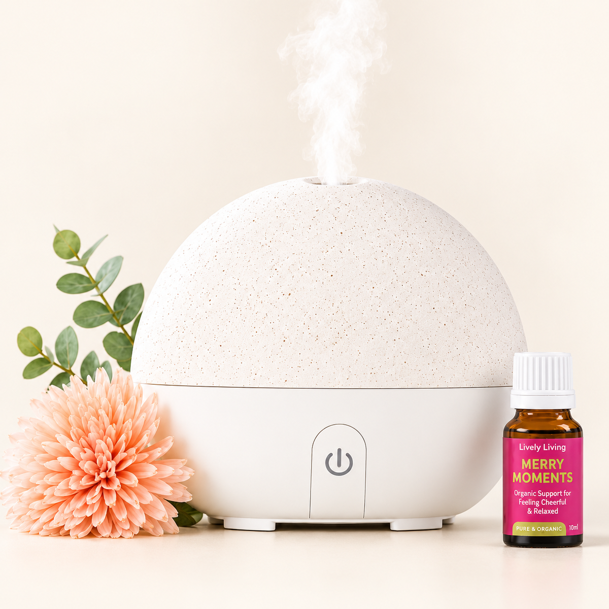 A round white and beige essential oil diffuser with mist coming from the top, styled with a peach flower, green leaves, and a small amber bottle labeled 'Lovely Living Merry Moments' on a warm cream background.