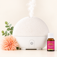 A round white and beige essential oil diffuser with mist coming from the top, styled with a peach flower, green leaves, and a small amber bottle labeled 'Lovely Living Merry Moments' on a warm cream background.