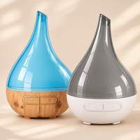 Two Aroma-Bloom teardrop diffusers, one translucent blue with a wood-look base and one translucent grey with a white base, in soft sunlight on a beige background