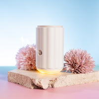 White Lively Living Aroma-Ascension waterless diffuser with a warm glow on a stone slab between pink pompom flowers