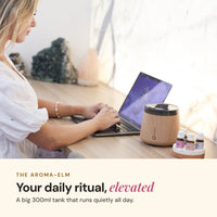 Woman working at a laptop beside a wood-look Aroma-Elm diffuser releasing mist, with small Lively Living oil bottles, headline Your daily ritual, elevated and a big 300ml tank that runs quietly all day