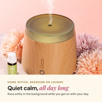 Wood-look Aroma-Elm diffuser misting beside a small Lively Living oil bottle and pink flowers, with the text Quiet calm, all day long for home office, bedroom or lounge