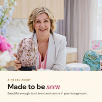 Smiling woman at a table with the speckled pink Aroma-Jewel Glass Diffuser, with the text Made to be seen: beautiful enough to sit front and centre in your lounge room