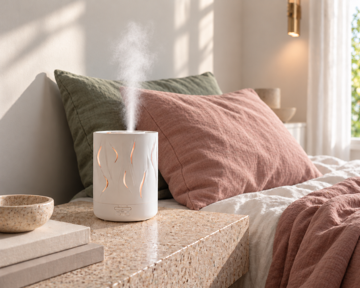 White Aroma-Cloud diffuser with wave cut-outs releasing mist on a stone bedside table in a warm bedroom with dusty pink and sage bedding