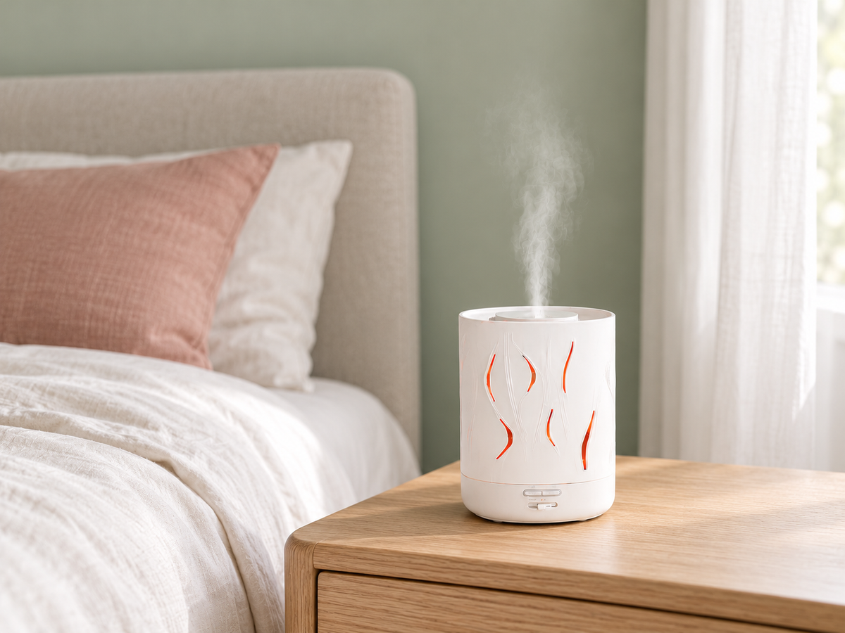 White Aroma-Cloud diffuser with wave cut-outs releasing mist on an oak bedside table beside a bed with pink and white linen