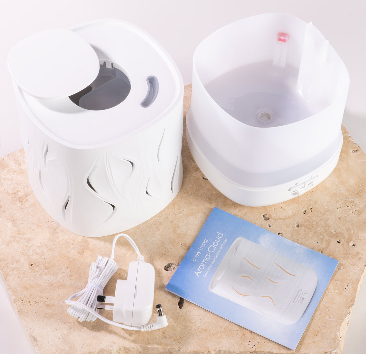 A white diffuser set shown on a light stone surface, including a cylindrical diffuser body with decorative cutouts, a separate white top section, a power adapter with cord, and a blue instruction card.