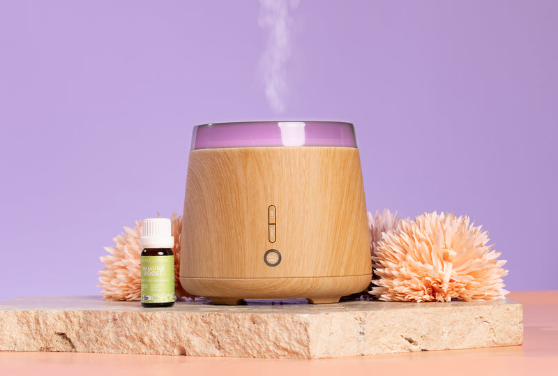 Wooden diffuser with essential oil bottle and flowers on a stone surface against a purple background