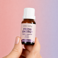 A hand holding a 15ml amber glass bottle of Lively Living Fresh Energy essential oil against a soft pink and purple gradient background.