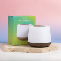 A white cylindrical aroma diffuser with a dark wood base stands in front of its green and blue packaging box on a light stone slab, set against a pink and blue gradient background.