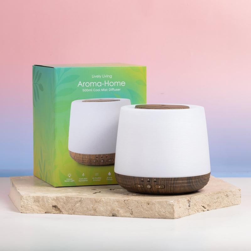 A white cylindrical aroma diffuser with a dark wood base stands in front of its green and blue packaging box on a light stone slab, set against a pink and blue gradient background.