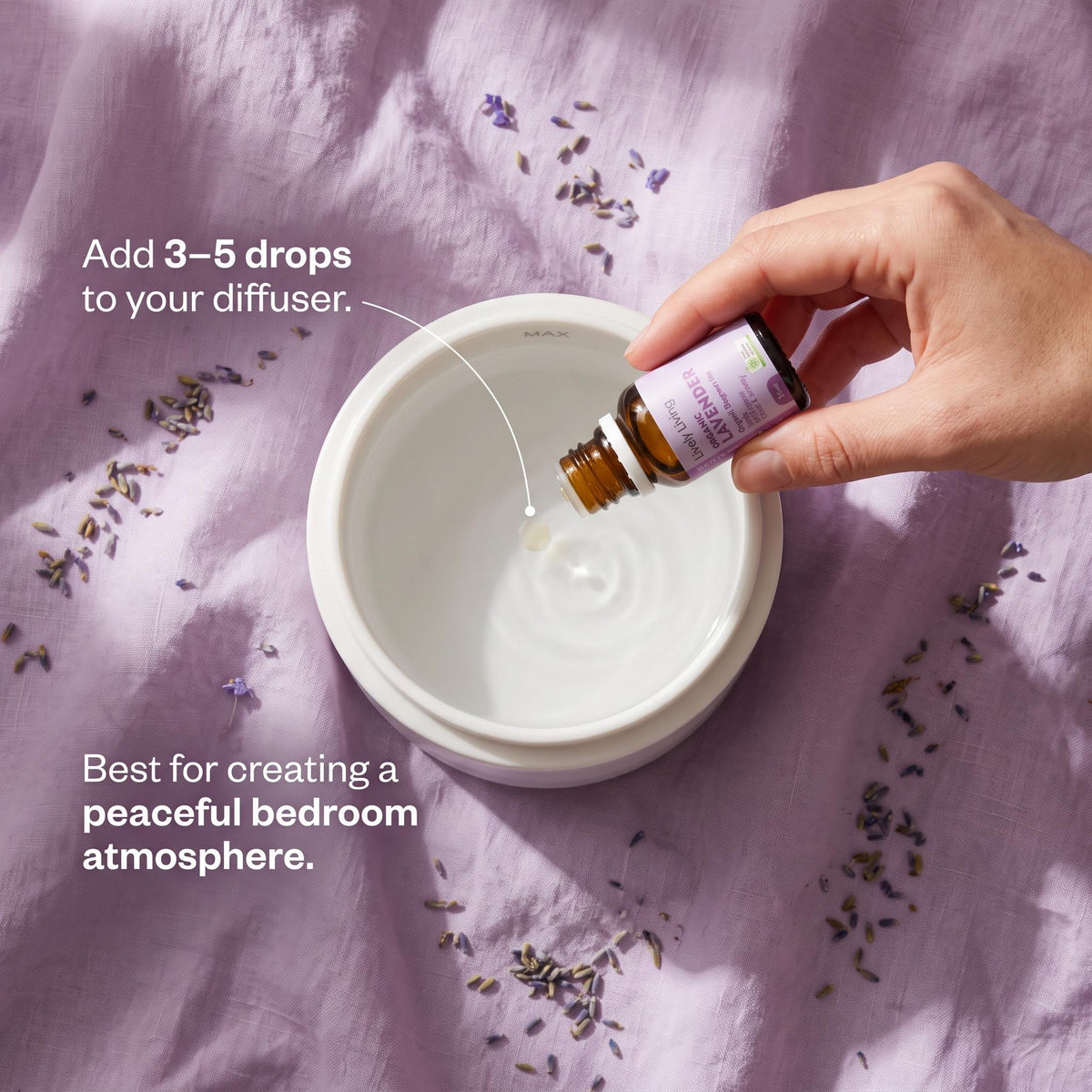 Overhead view of a hand adding drops of Lively Living Organic Lavender oil to a white diffuser on lilac linen scattered with dried lavender buds, with the text Add 3-5 drops to your diffuser