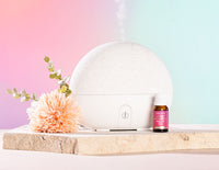 White diffuser with flowers and a bottle on a wooden surface against a pastel gradient background