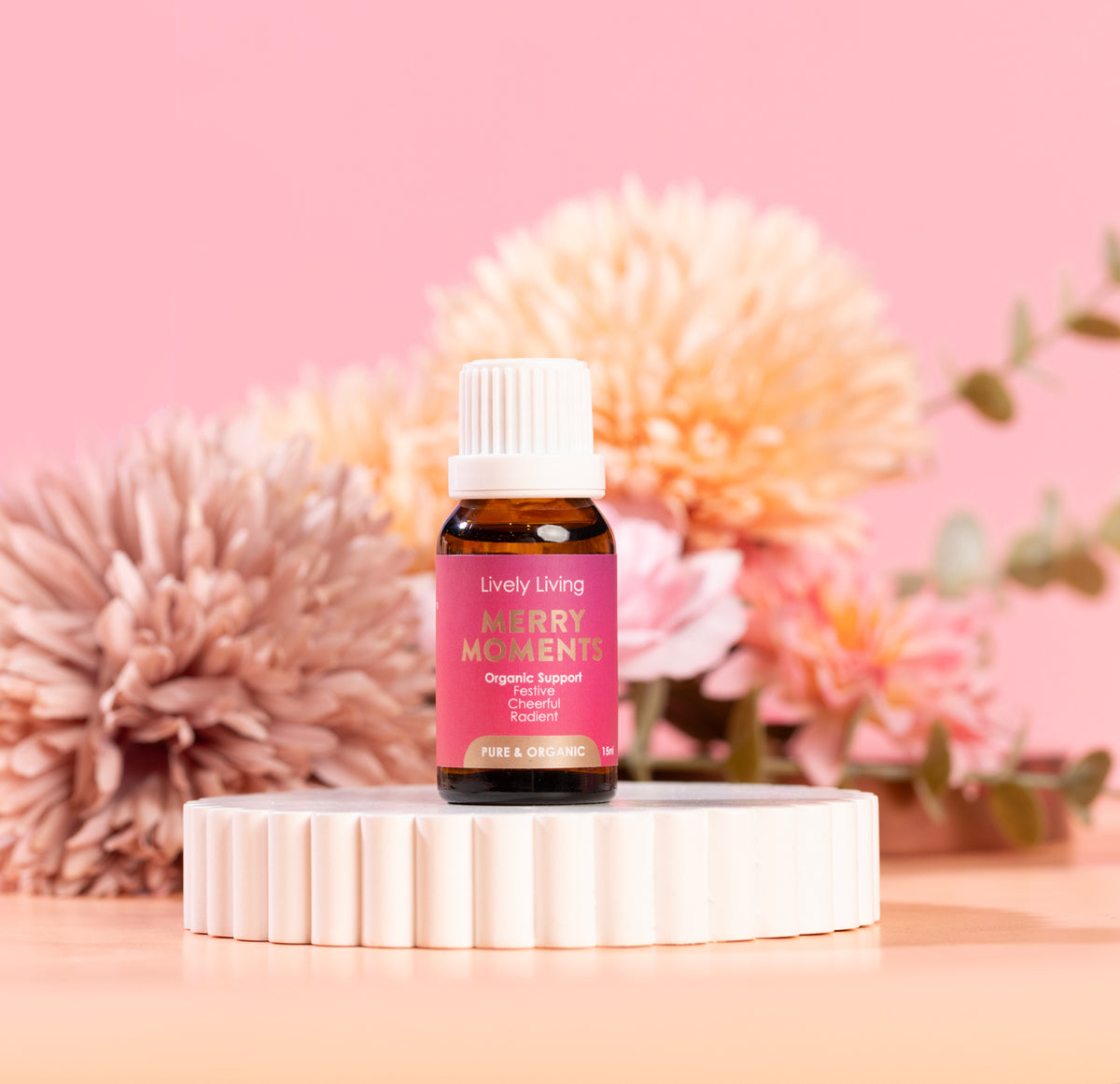 Bottle of Lively Living Merry Moments essential oil on a white stand with pink flowers in the background