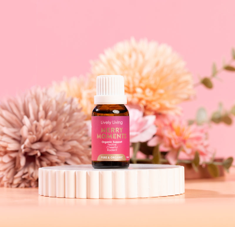 Bottle of Lively Living Merry Moments essential oil on a white stand with pink flowers in the background