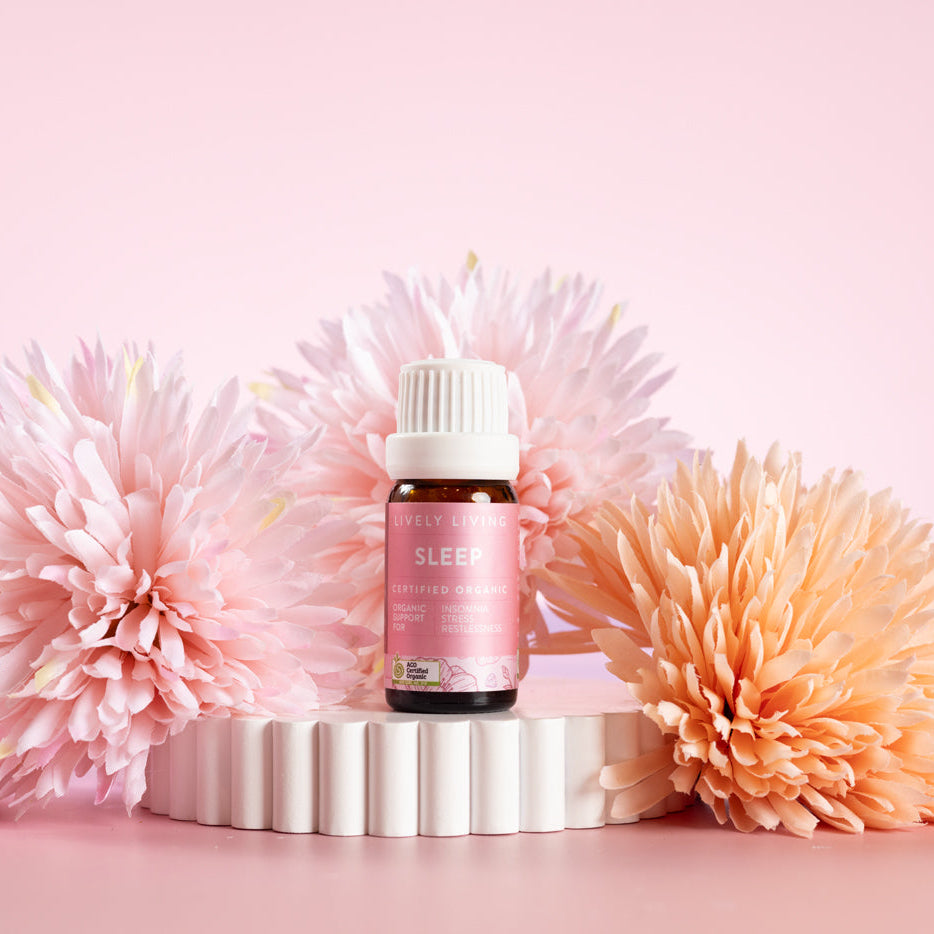 Lively Living Certified Organic Sleep Essential Oil Blend. Sitting on a rippled white plinth surrounded by pink and orange flowers. 