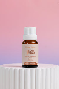 A small amber glass bottle with a white ribbed cap and a beige label reading 'Lively Living Flow Living' stands on a white ribbed pedestal against a soft pink-to-lavender gradient background.