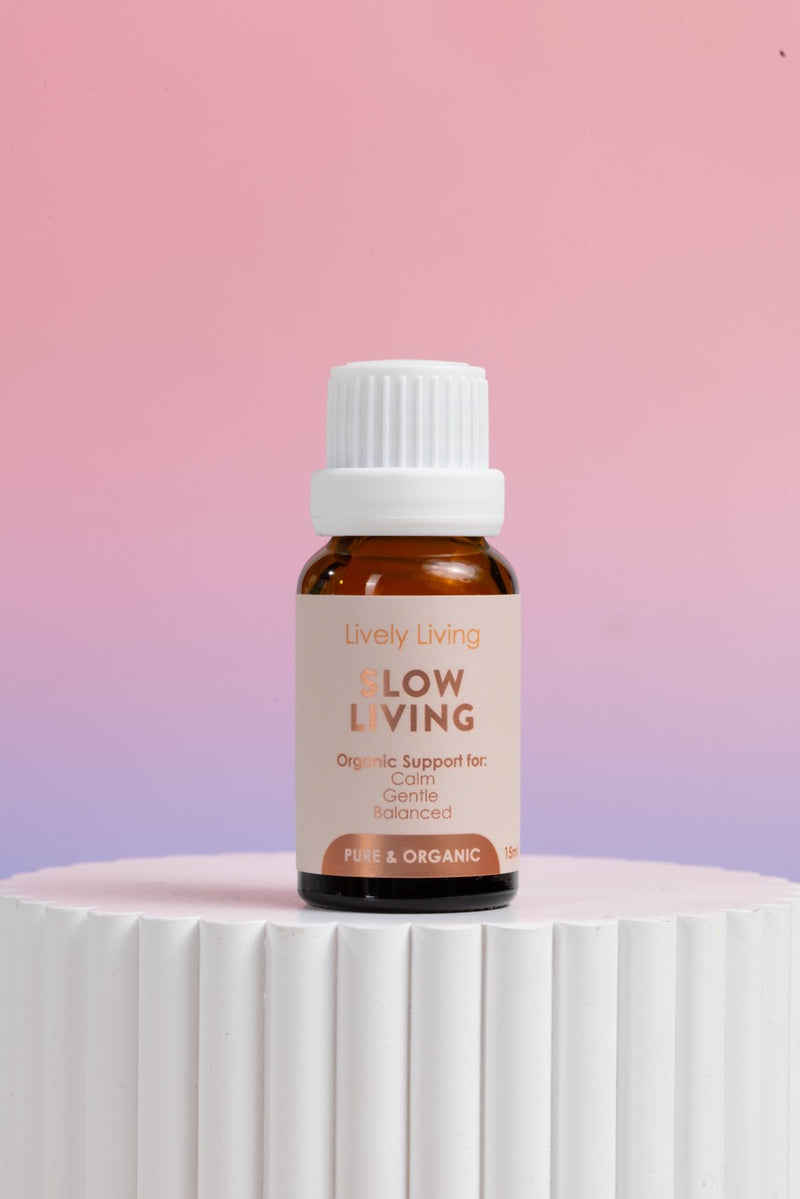 A small amber glass bottle with a white ribbed cap and a beige label reading 'Lively Living Flow Living' stands on a white ribbed pedestal against a soft pink-to-lavender gradient background.