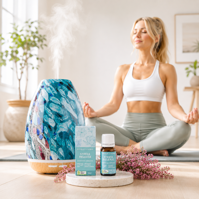 Woman meditating with a diffuser, flowers, and bottles on a table in a bright room.