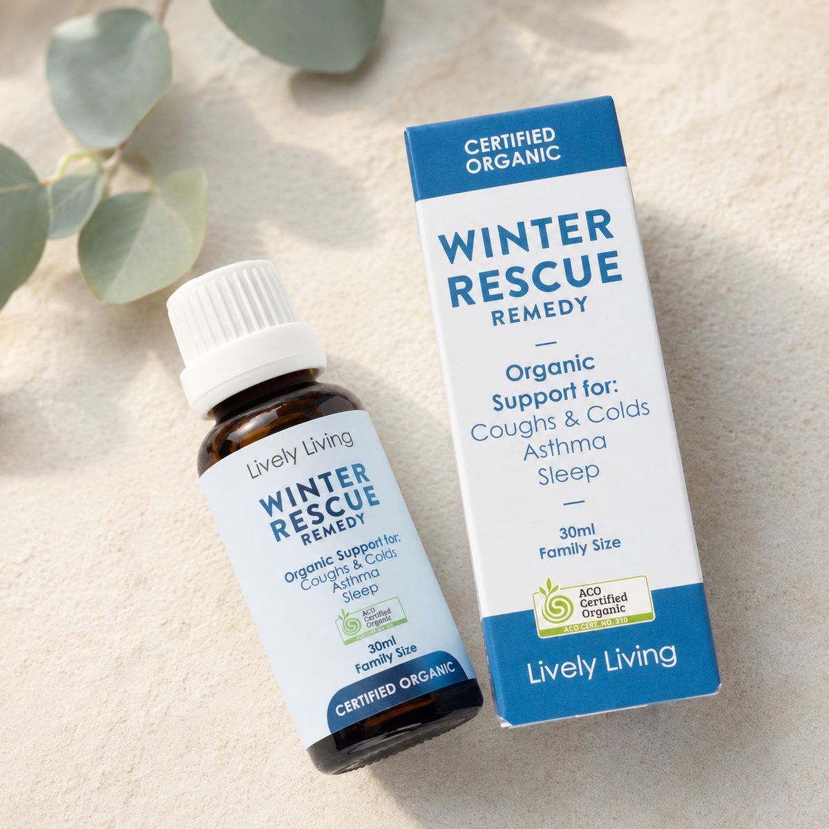 A small amber bottle and matching box of Lively Living Winter Rescue Remedy, photographed on a light beige textured surface with soft green leaves in the background.