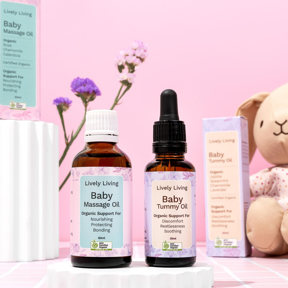Baby Massage Oil and Baby Tummy Oil with boxes on white pedestals beside a plush bunny and purple flowers